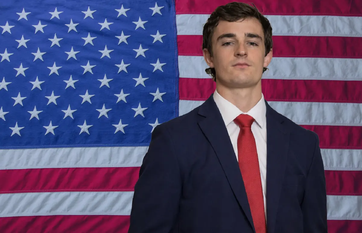 Nick Shirley in a navy suit and red tie in front of an American flag